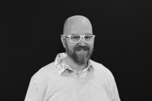 A black and white headshot of board member Wes Harris. He is wearing a light-coloured shirt and light-coloured statement glasses. The background is black.