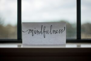 The word mindfulness written in cursive in black ink on white paper rests on a windowsill. The window has black panes and the sky appears cloudy outside.