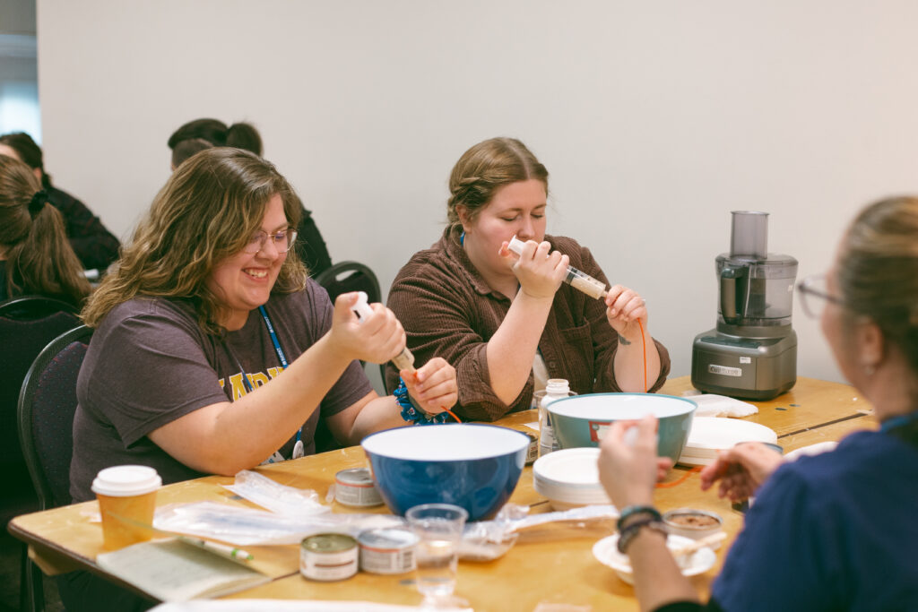Two Registered Veterinary Technicians participate in a workshop, practicing tube feeding techniques. They are seated at a table with two bowls and other equipment in front of them.