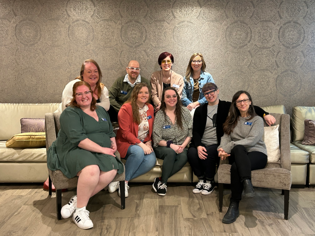 The nine members of the OAVT Board of Directors sit on a sofa and two wing chairs with three people standing behind the sofa.