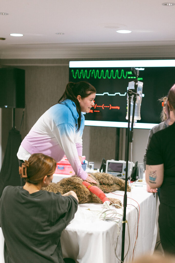 A woman performs CPR on a dog mannequin. A woman in the foreground holds the mannequin's head while a third woman, to the right of the image, monitors vitals projected on a large screen.